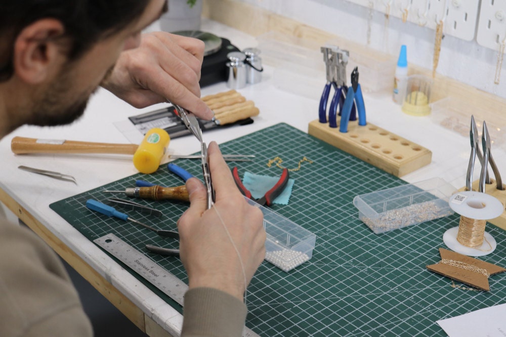 Sabrina's partner uses pliers to assemble a dainty chain at their jeweler's workbench.