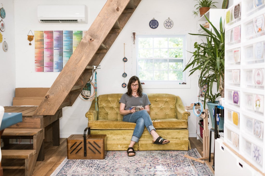 Liz sits on a couch in her cozy studio space and works on some embroidery