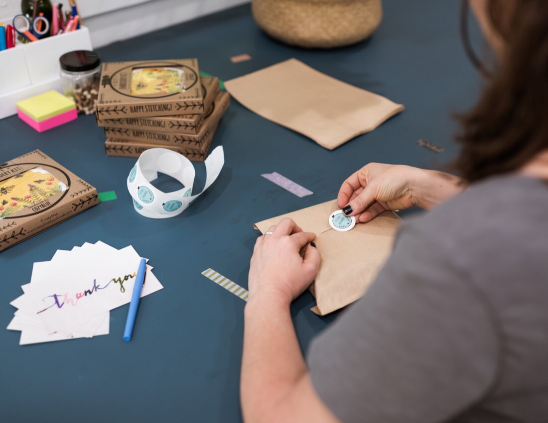 Liz packages an embroidery hoop kit for shipment, adding a Cozy Blue sticker to the outside