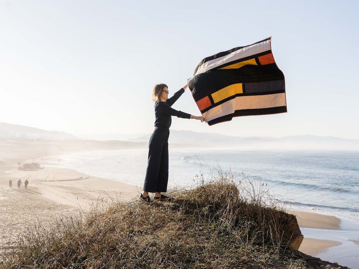 A woman overlooking a beach holds up a color-blocked quilt from Vacilando Quilting, letting it float in the wind