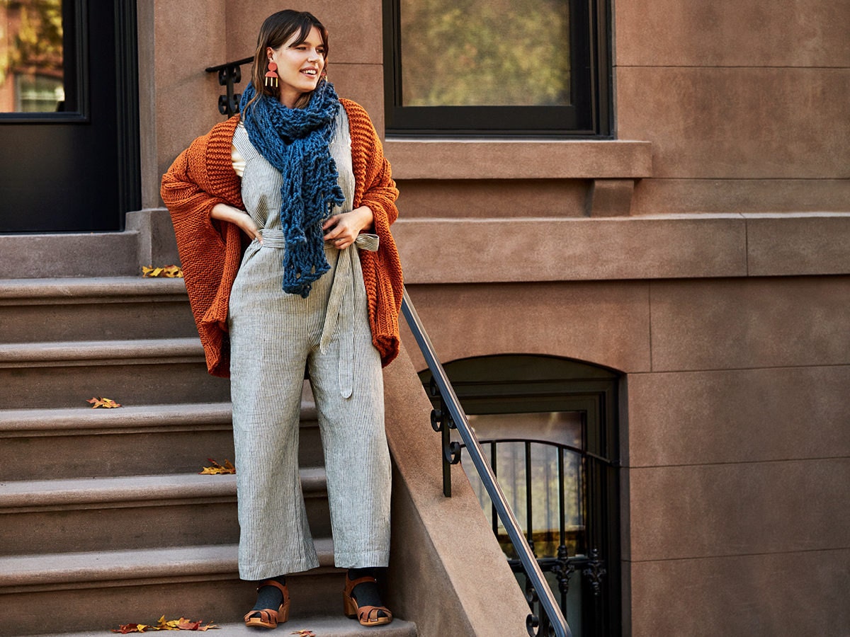 Woman standing on stoop stairs wearing a linen jumpsuit, knit cardigan, oversized scarf, and clogs