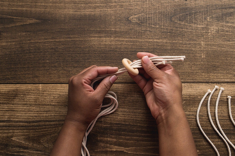 Hands pull four cotton macrame ropes through a wooden hoop.