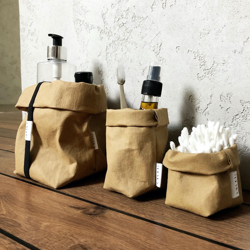 A trio of washable paper market bags in various sizes, shown storing bathroom staples like cotton swabs and toothbrushes.