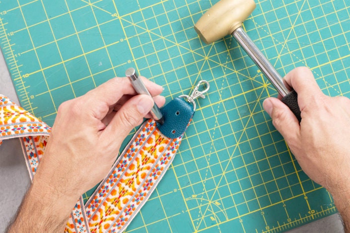 A mallet and leather hole punch are used to punch three holes through all the layers of the folded leather and trim near each end of the strap