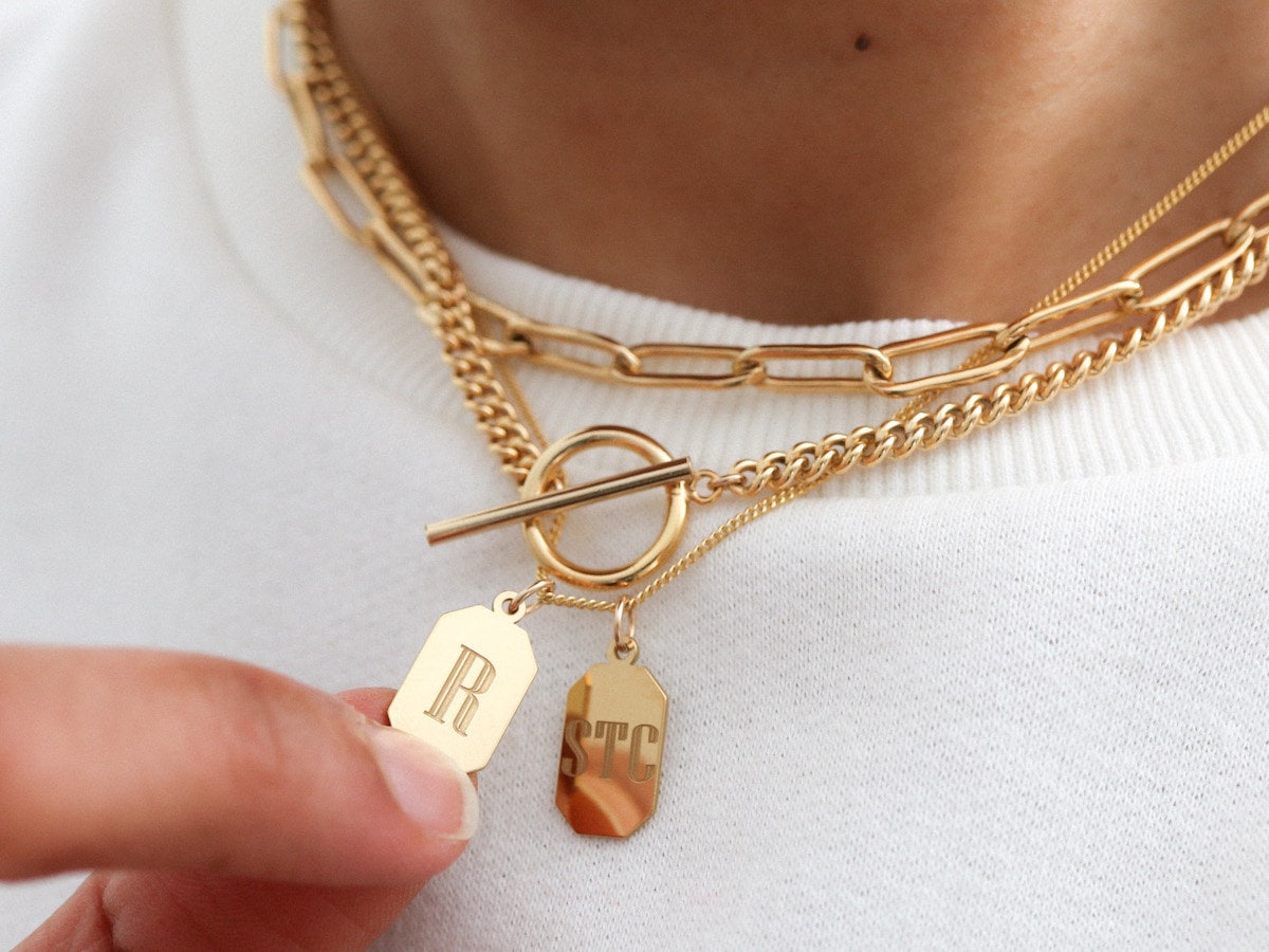 A gold engraved initial pendant necklace layered over gold chains from EVREN