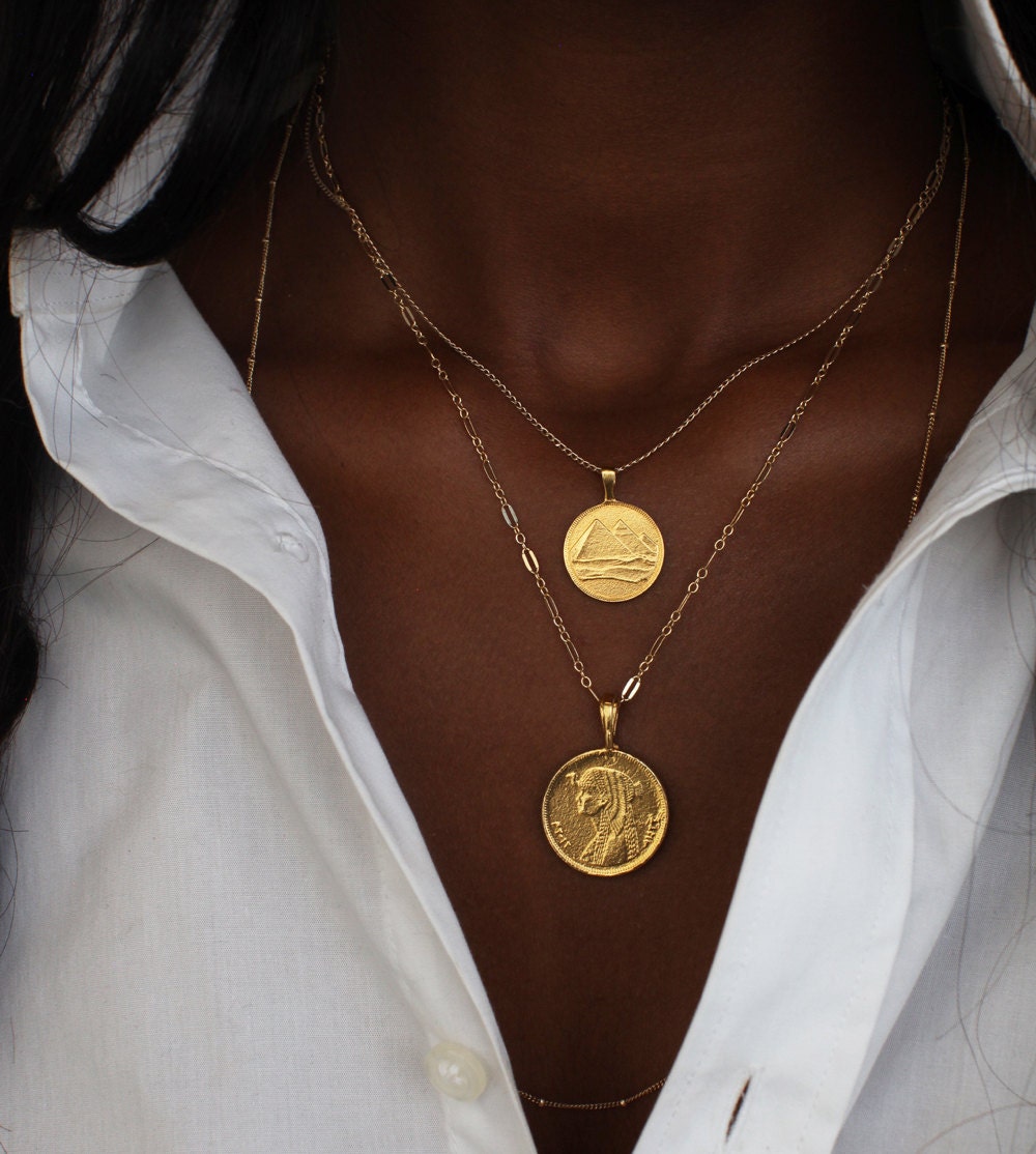 A woman models a stack of two necklaces from Omi Woods: a pyramid charm necklace and a Cleopatra coin pendant necklace