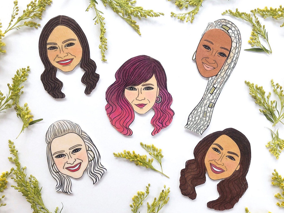 Five custom-drawn face magnets on a white surface with flowers.