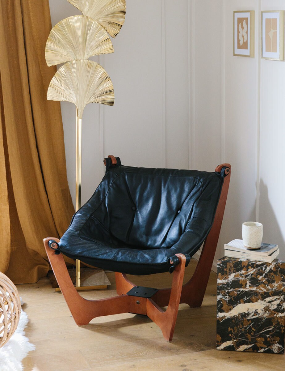 A black leather sling chair sits next to a gold ginkgo leaf lamp and a stone side table with books and a coffee cup on top.