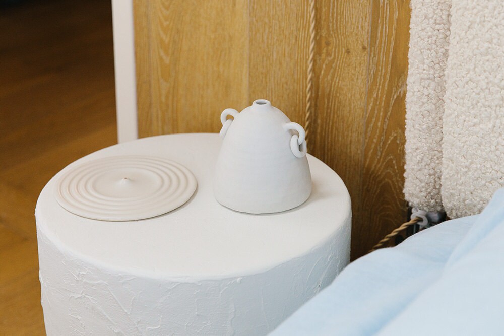 A cream-colored rippled incense holder and a matte ceramic vase rest atop a white plaster side table.