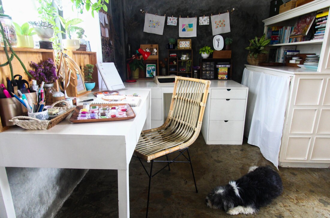 A pup lies at the foot of Ruby's work space, which is full of colorful artwork, crafting books, and embroidery supplies
