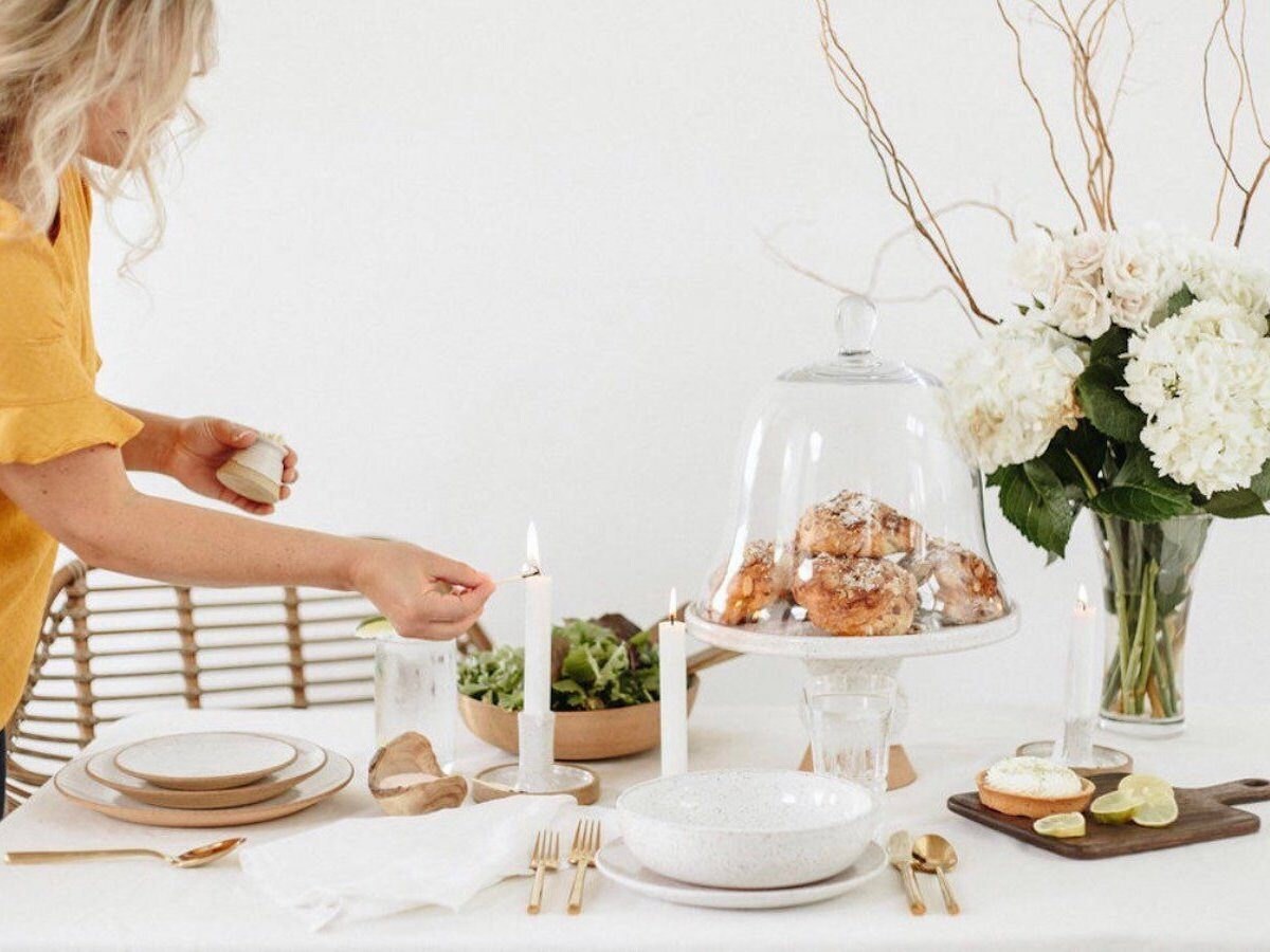 A woman lights a candle atop a set dining table, while holding a match holder with built-in striker by Blanc Pottery