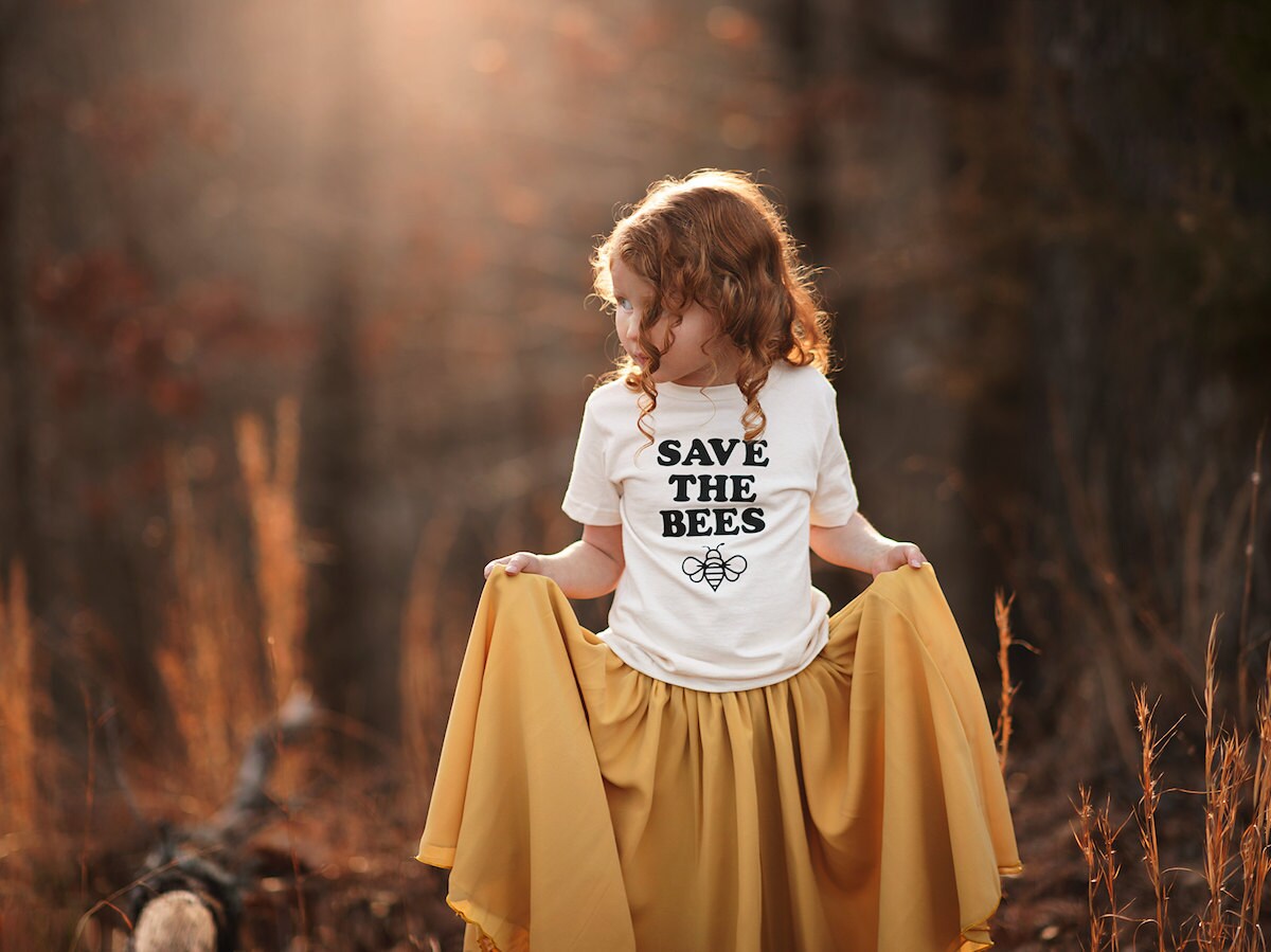 A little girl in a field wearing a "Save The Bees" T-shirt from Nature Supply Co.