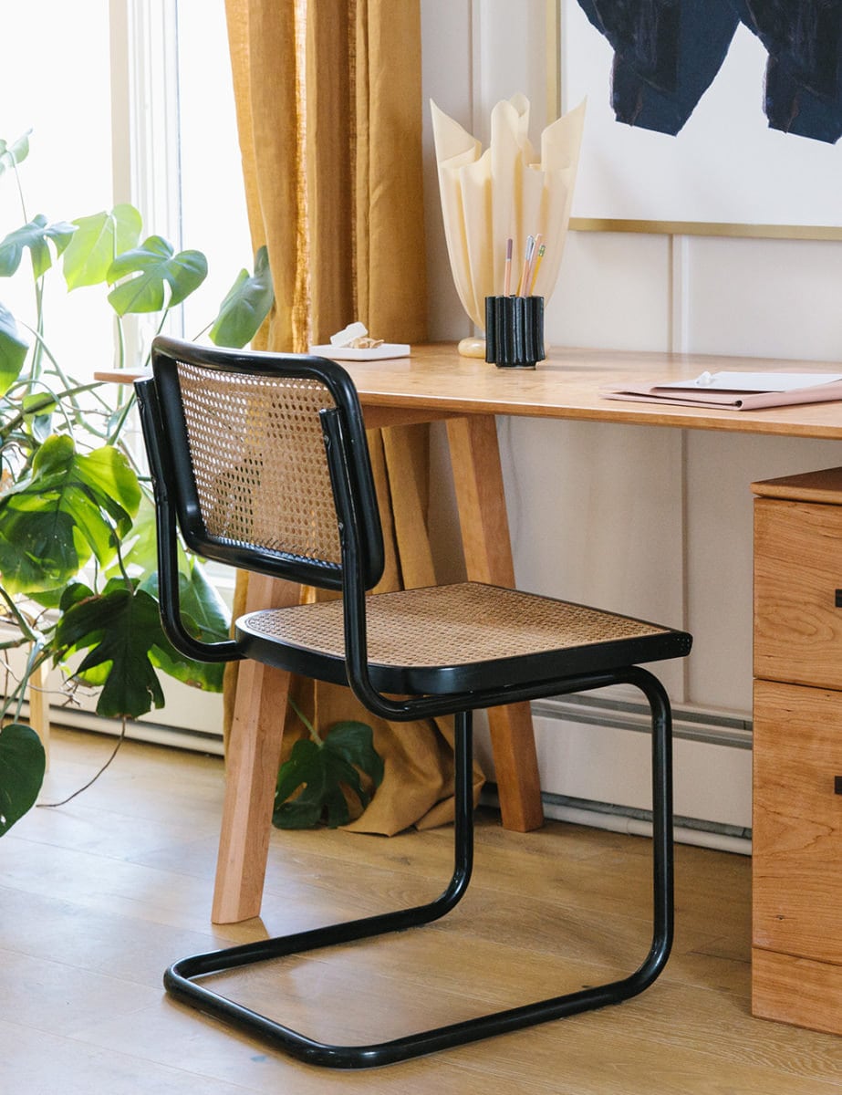 A black rattan Cesca chair sits in front of a wooden desk. Nearby is a leafy green Monstera plant.