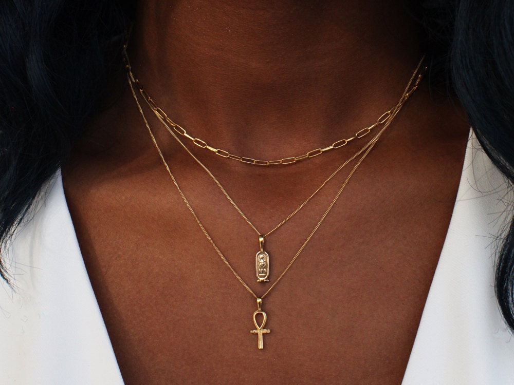 A woman models a stack of necklaces from Omi Woods, including a cartouche necklace and an ankh necklace.