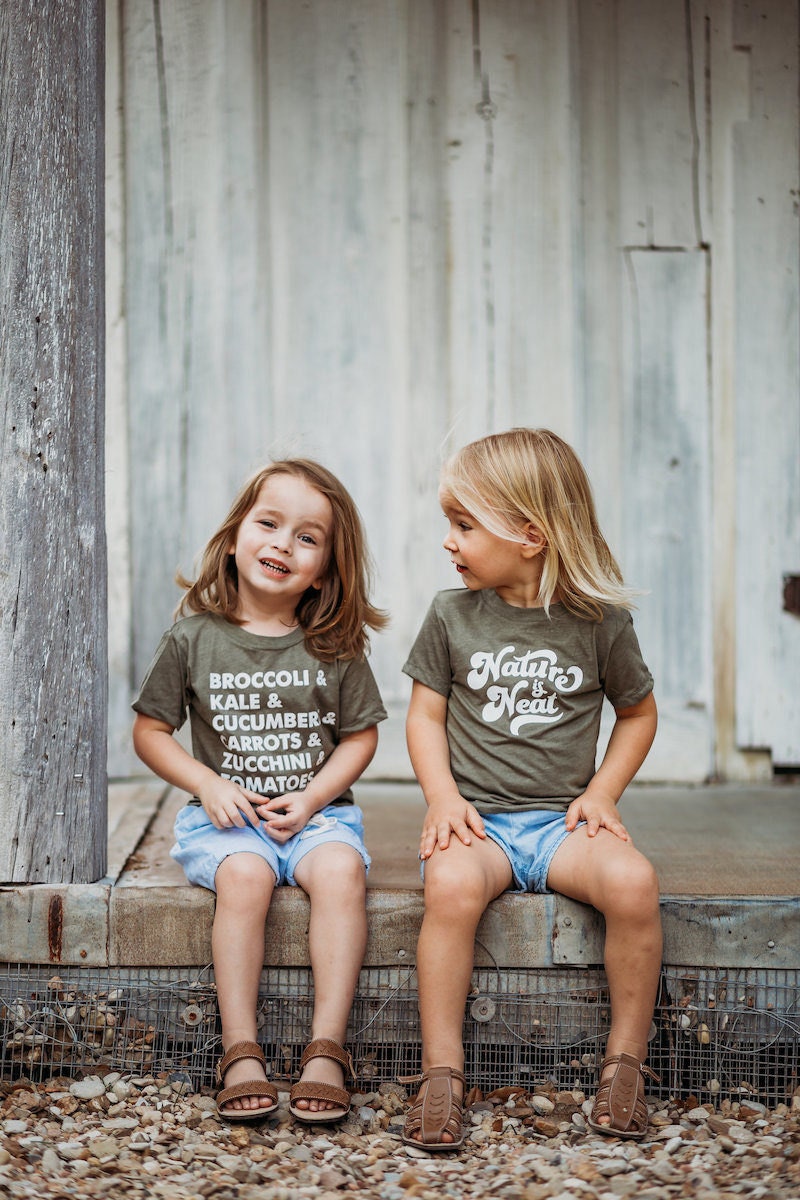 Two children sit wearing olive green T-shirts from Nature Supply Co.