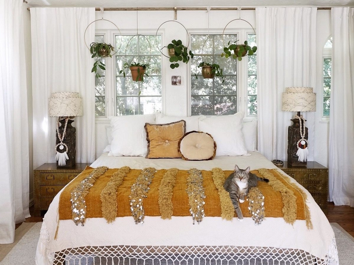 A bright bedroom interior featuring hanging planters, a bed covered with an orange blanket and framed by two nightstands, and a cat