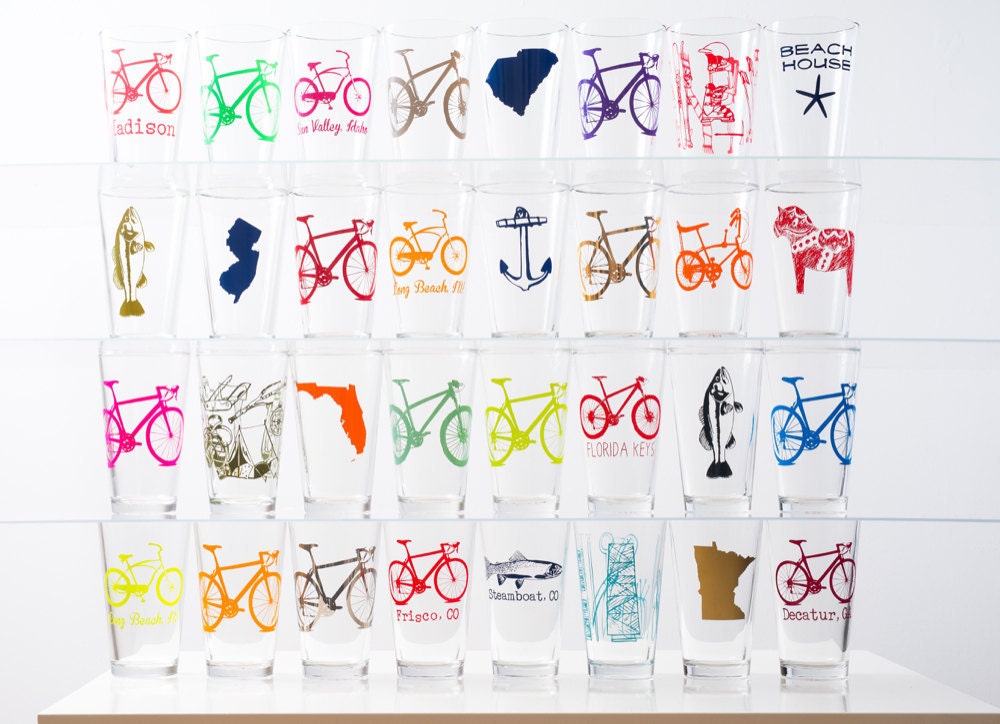 Four rows of neatly stacked pint glasses featuring an assortment of colorful printed illustrations, from fish and bicycles to horses and states.