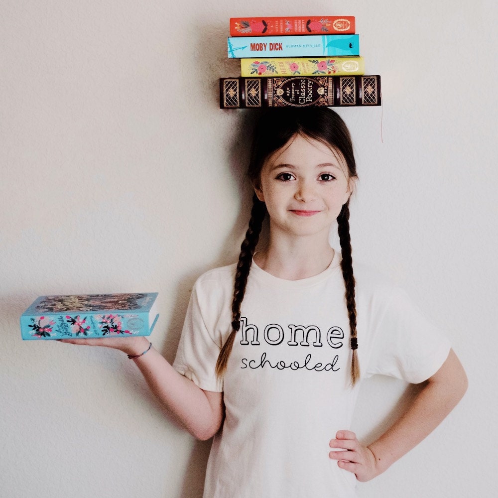 A girl wearing a "Homeschooled" T-shirt from Nature Supply Co., with a stack of books balanced on her head