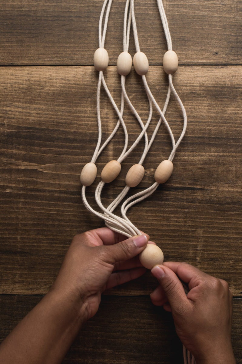 Hands pull all eight ropes through the bottom bead of the DIY macrame-inspired plant hanger.