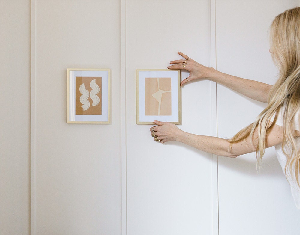 Sarah hangs a small framed art print on the wall, next to another similar design.