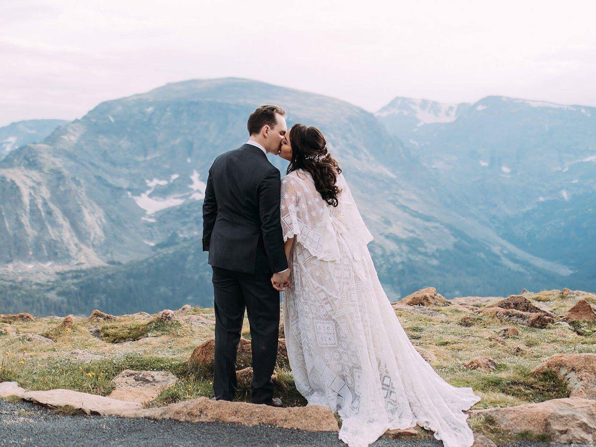 A bride wearing a wedding dress by Reclamation kisses her groom in front of a scenic mountain backdrop.