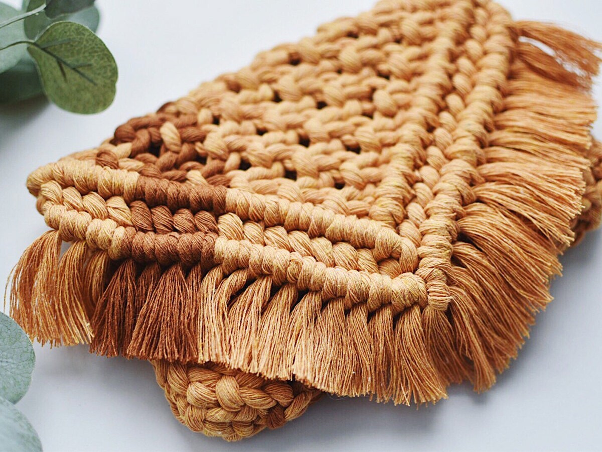 A beige macramé cutch with tassels on a white background with greenery.