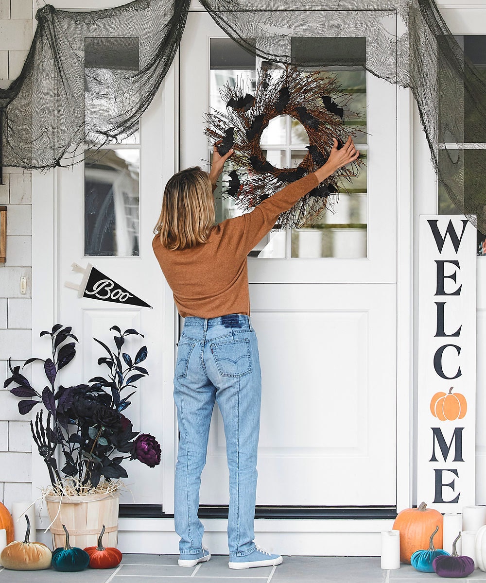 A woman hangs a sparkly bat wreath on the front door of a porch decorated for Halloween