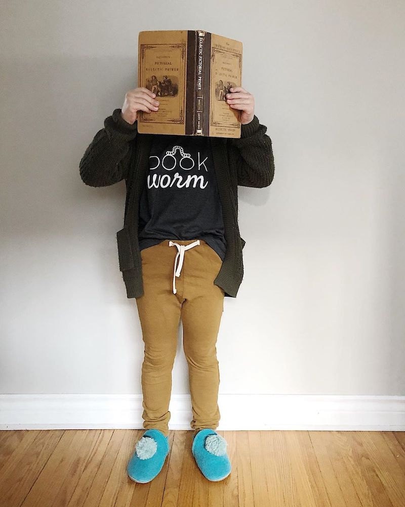 A child wearing a "Bookworm" T-shirt from Nature Supply Co. holds a book up to his face