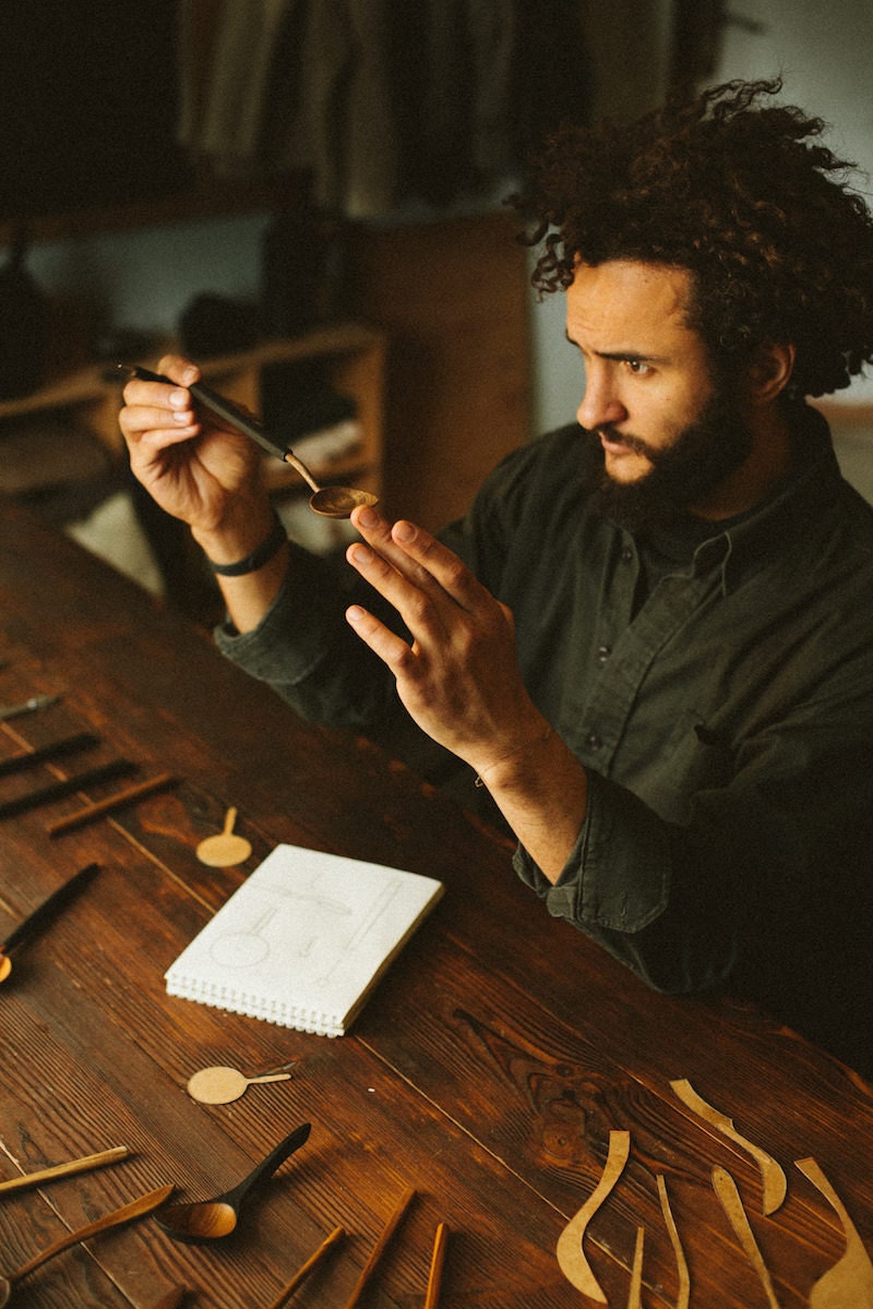 Kirill sits at a table, surrounded by a sketchbook and several finishes pieces, and inspects a wooden spoon