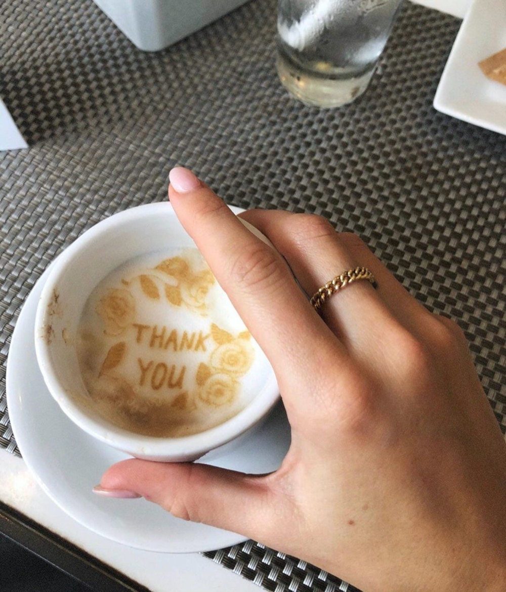 A hand wearing a gold chain stacking ring holds a coffee cup with the words "thank you" written in the foam.