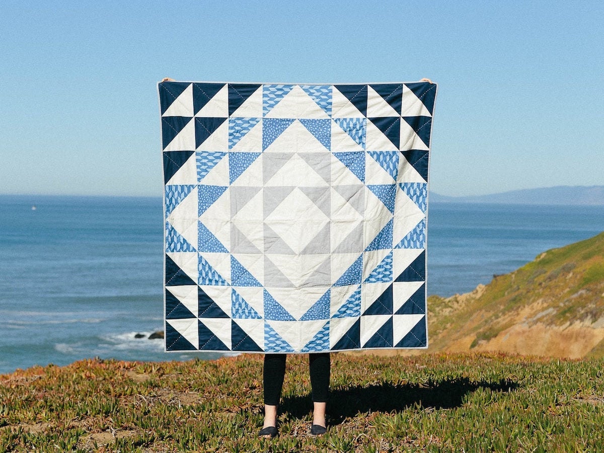 A blue and white quilt held up in front of the ocean