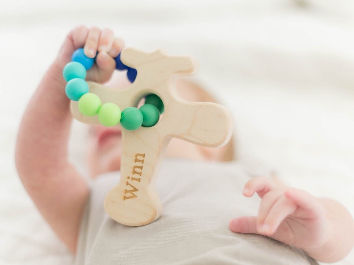 A baby holds a personalized wooden teether shaped like an airplane with blue and green silicone beads