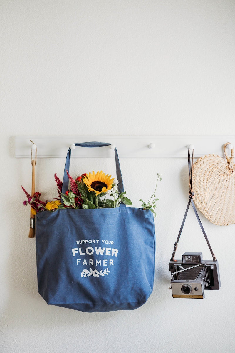 A reusable blue tote bag from Nature Supply Co. hangs on a peg board filled with flowers