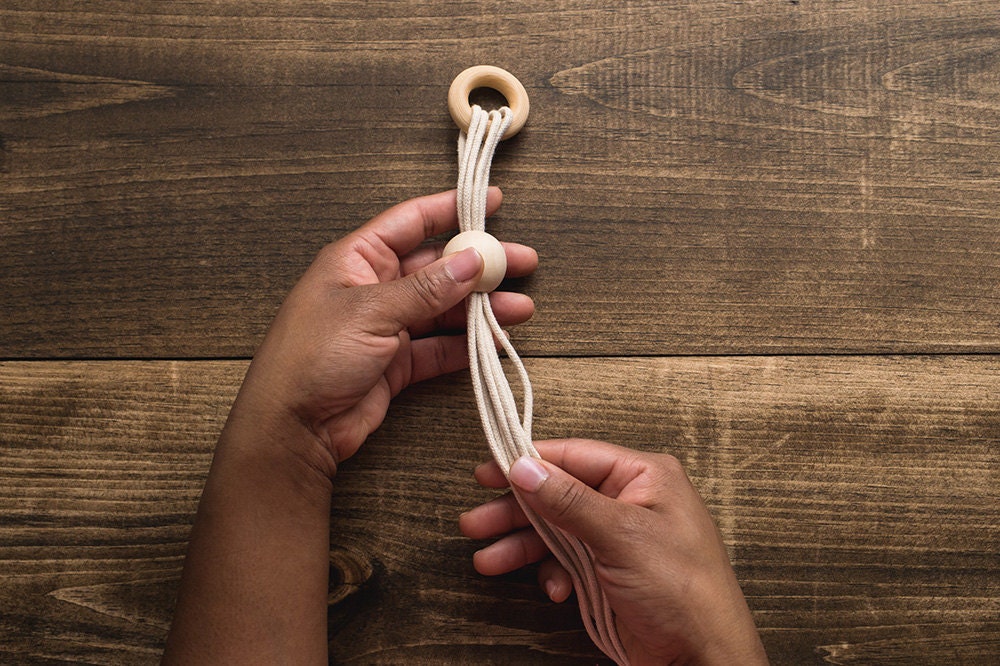 Hands add a bead to the top of the DIY macrame-inspired plant hanger.