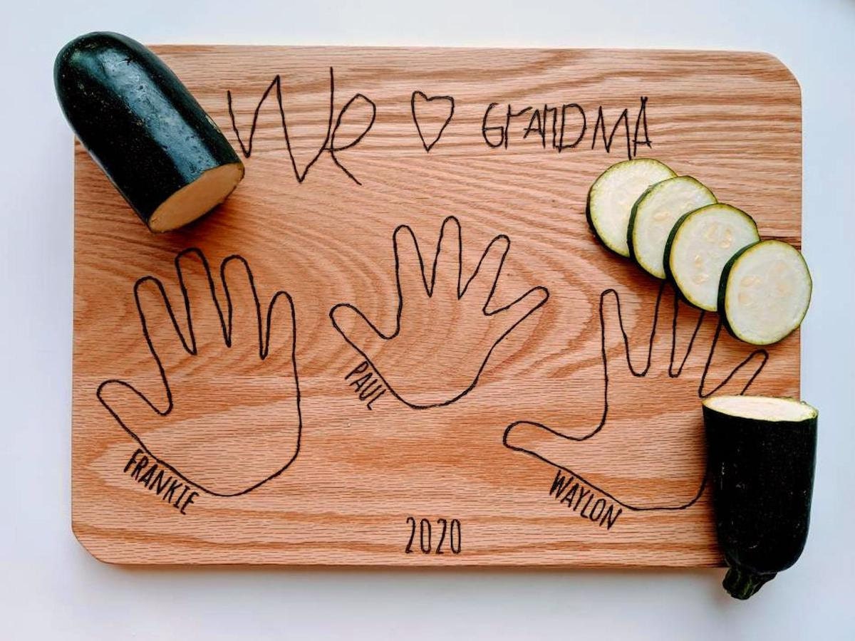 A personalized cutting board engraved as a gift for grandma, with the outlines of the handprints of 3 grandchildren.