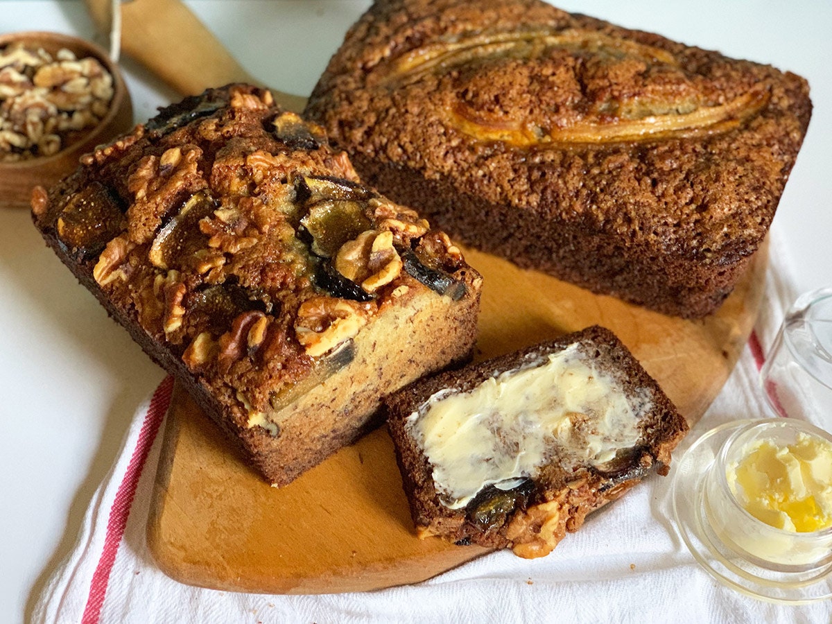 Two loaves of freshly baked banana bread, one with a fig and walnut topping, and one with a sliced banana topping.