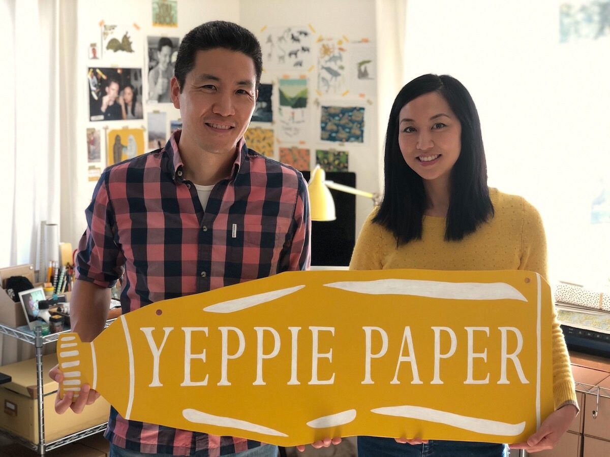 Jen and Jim standing in their studio holding a Yeppie Paper sign