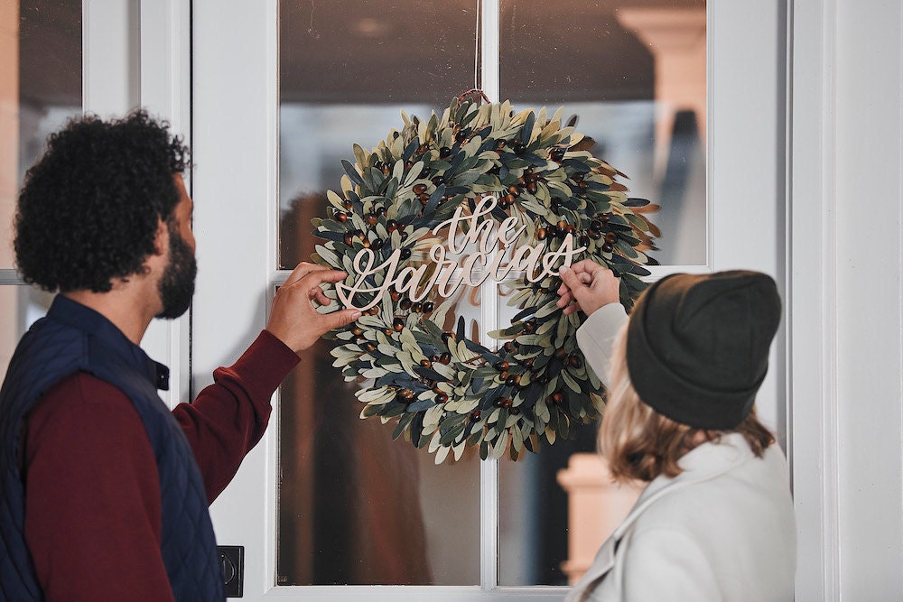 A couple hangs a faux olive wreath and calligraphy name sign that says "The Garcias" on their front door