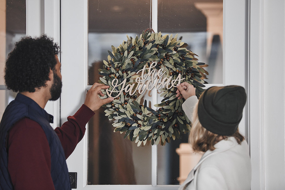 A couple hangs a faux olive wreath and calligraphy name sign that says "The Garcias" on their front door