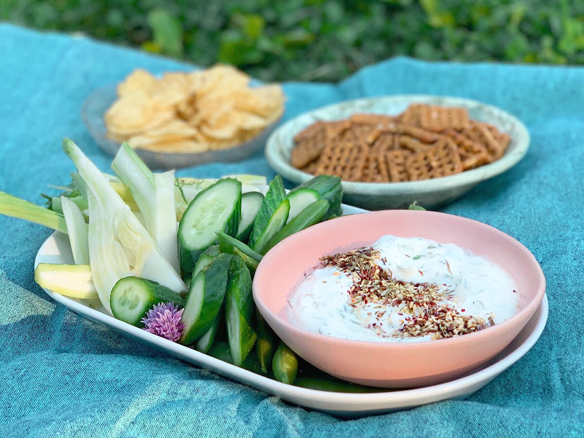 A styled picture of creamy yogurt herb dip on a picnic blanket, with sides