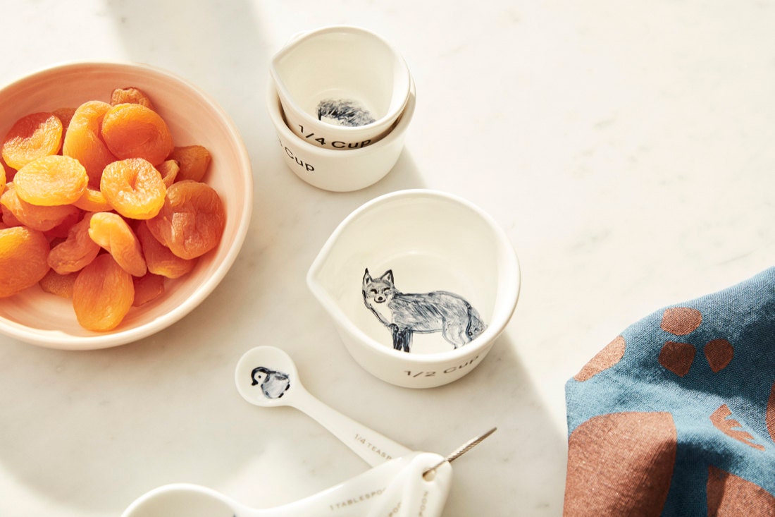 Hand-painted woodland animal themed measuring cups on a tabletop next to penguin measuring spoons