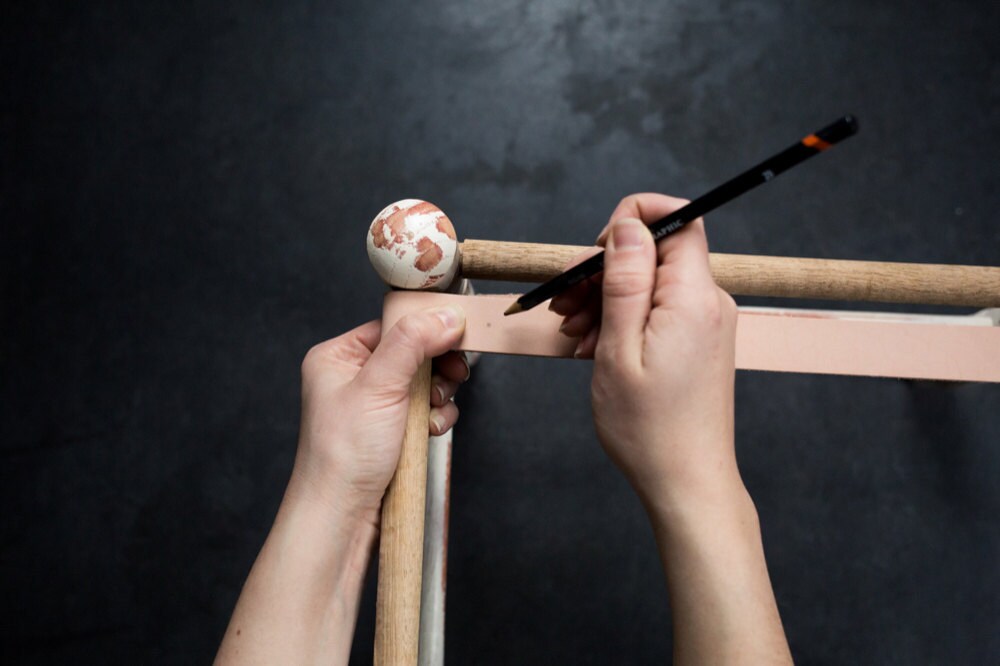 Using a pencil to mark a dot in the center of the overlapping section of the leather strap that's wrapped around the stool's frame for screw placement