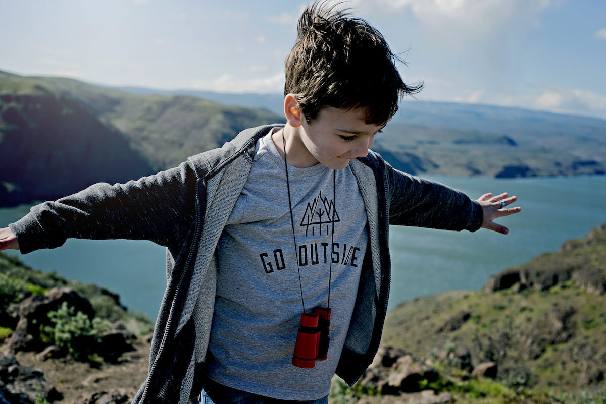 A boy with binoculars around his neck wearing a "Go Outside" T-shirt on a mountaintop