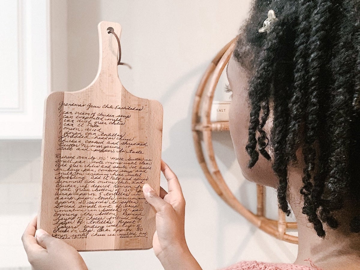 A woman holds up a wooden cutting board inscribed with a handwritten recipe.