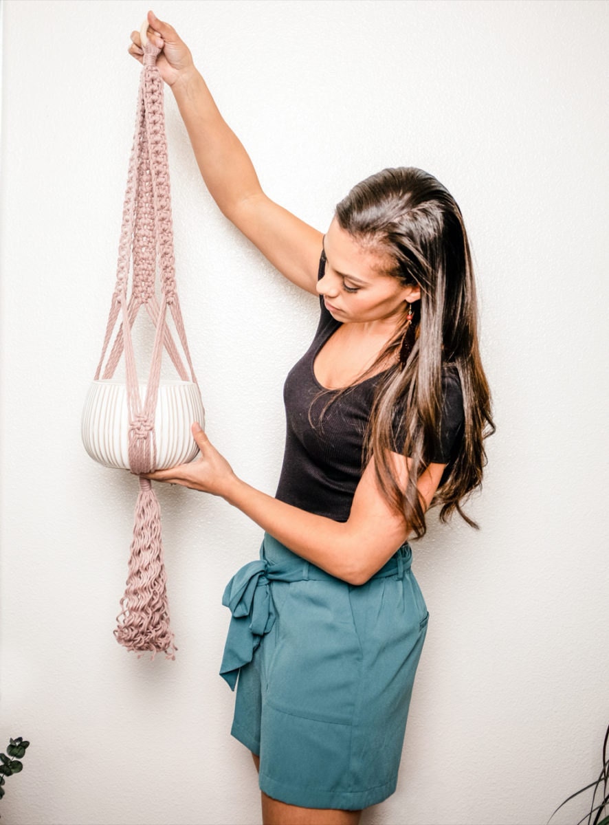 Casey holds up a blush-colored macrame plant hanger with curly fringe.