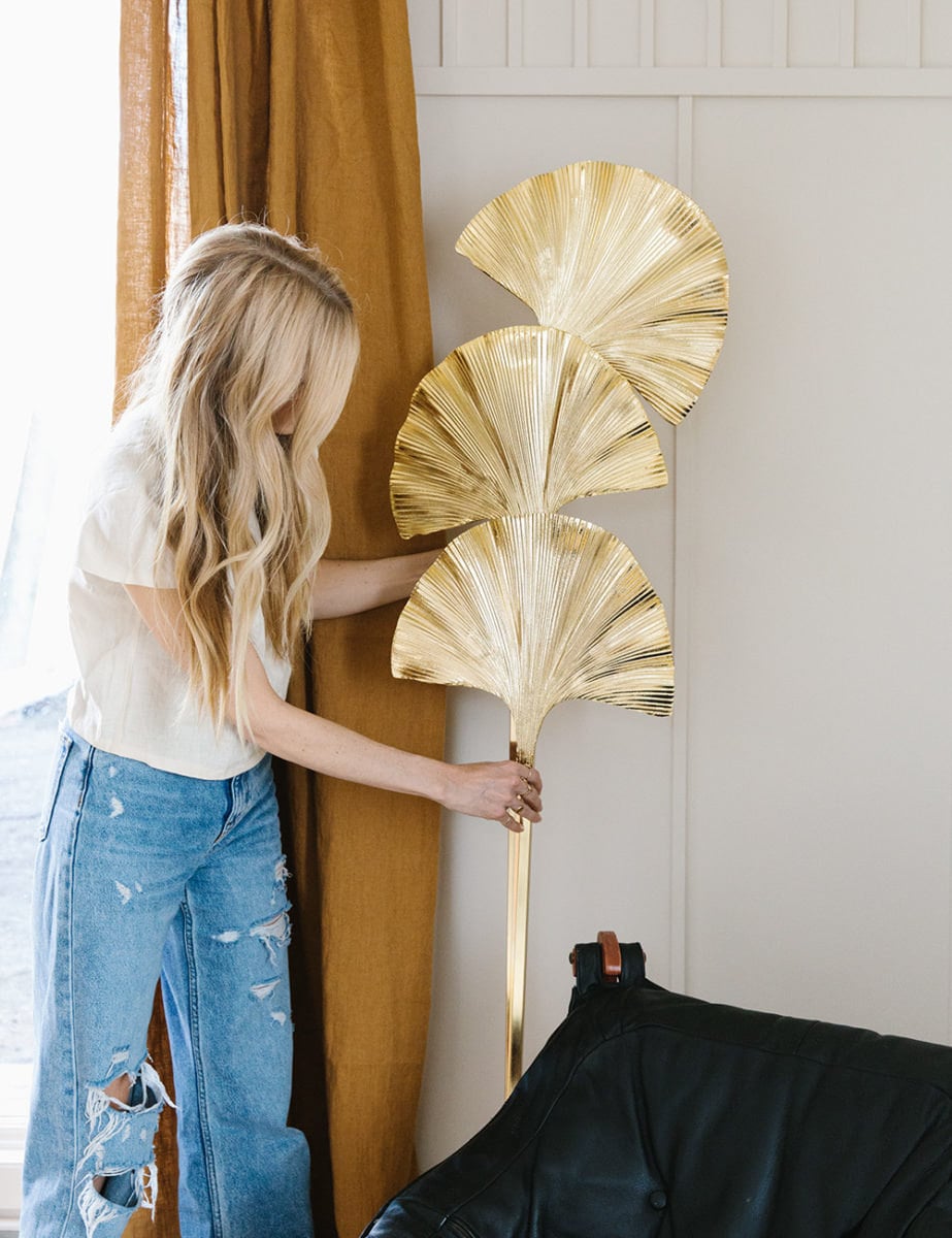 Sarah moves a gold standing floor lamp that is taller than her and has three ginkgo-leaf shapes.