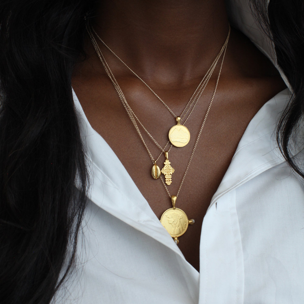 A woman models a stack of necklaces from Omi Woods, including her pyramid charm necklace, Ethiopian coptic cross, cowrie necklace, and Cleopatra coin necklace with pearls