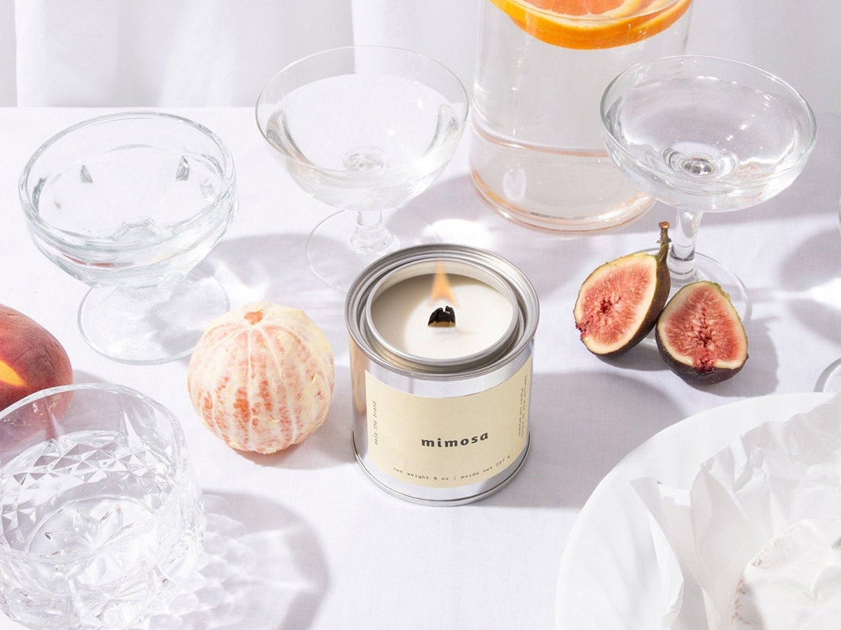 A silver mimosa-scented candle surrounded by glassware and fruit.