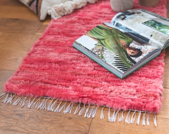 May include: A pink fluffy rug with a white fringe. The rug is on a wooden floor and a magazine is lying on top of it.