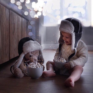 May include: A child wearing a brown and white fur-lined aviator hat sits on the floor with a teddy bear wearing a similar hat. Both are holding mugs filled with marshmallows.